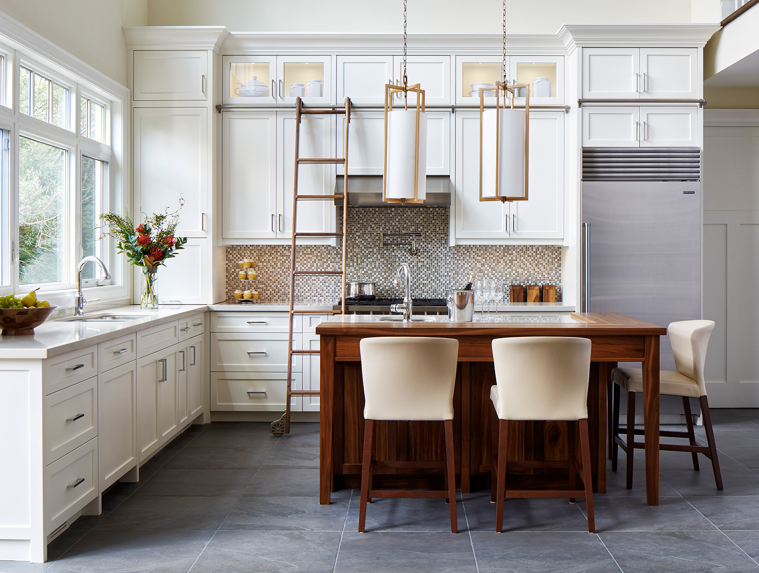 A photo of the project kitchen with white cabinets, grey floor tiles, a dark wood island, and a library ladder.