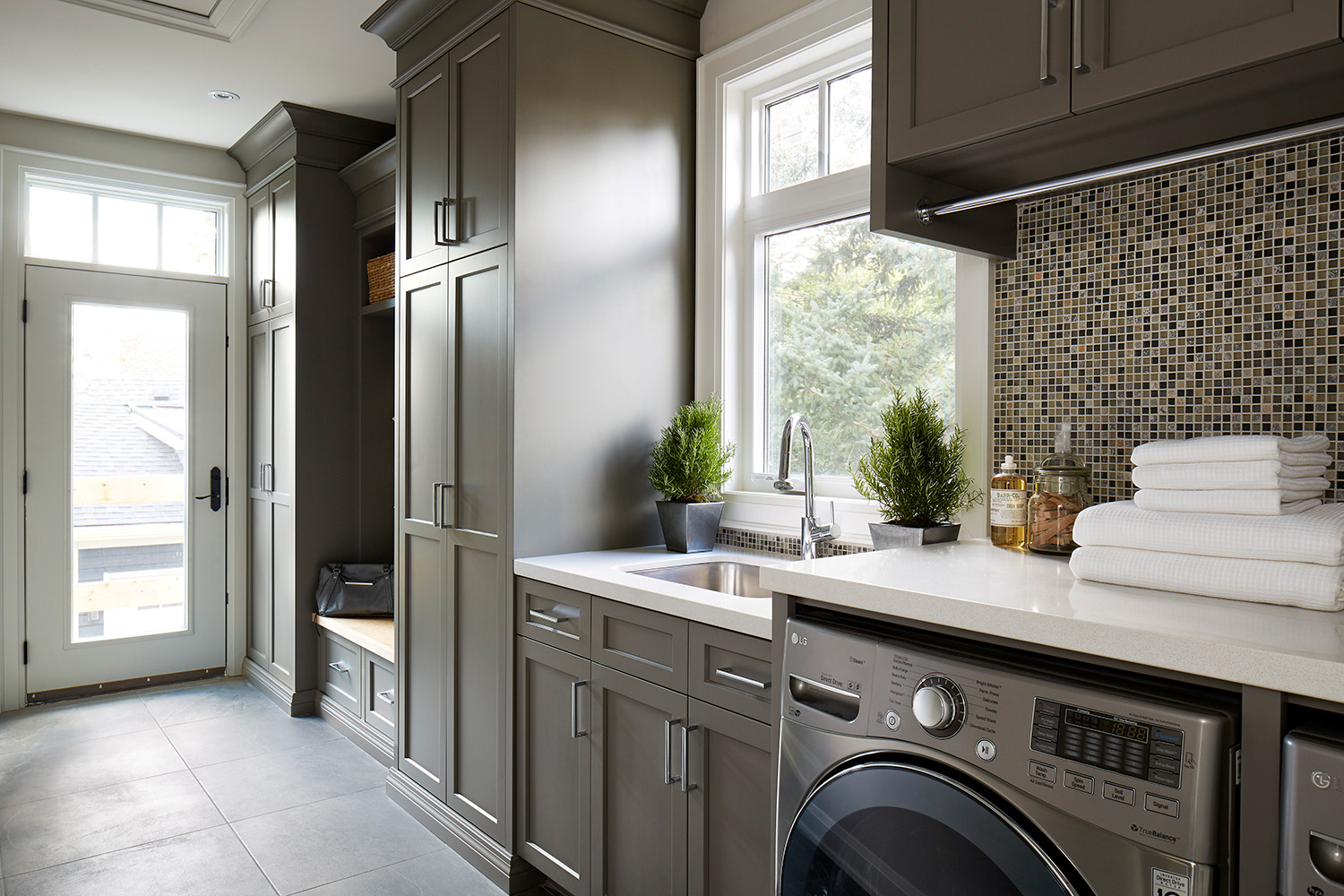 The project laundry room with dark olive green cabinetry, mosaic backsplash, stainless steel laundry appliances, and the laundry sink.