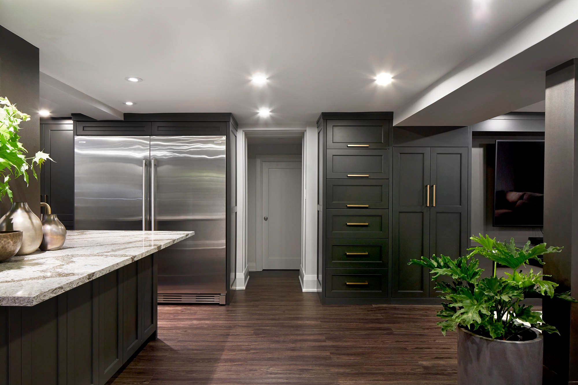 NUITH – FIFTY SHADES OF DIOR GRAY – BASEMENT MANCAVE STORAGE DESIGNED BY CELIA ALIDA RUTTE – PHOTOS BY DONNA GRIFFITH Dark grey custom cabinetry with brass handles, a dark walnut flooring, a large double sided fridge freezer, and a green plant in a grey pot.
