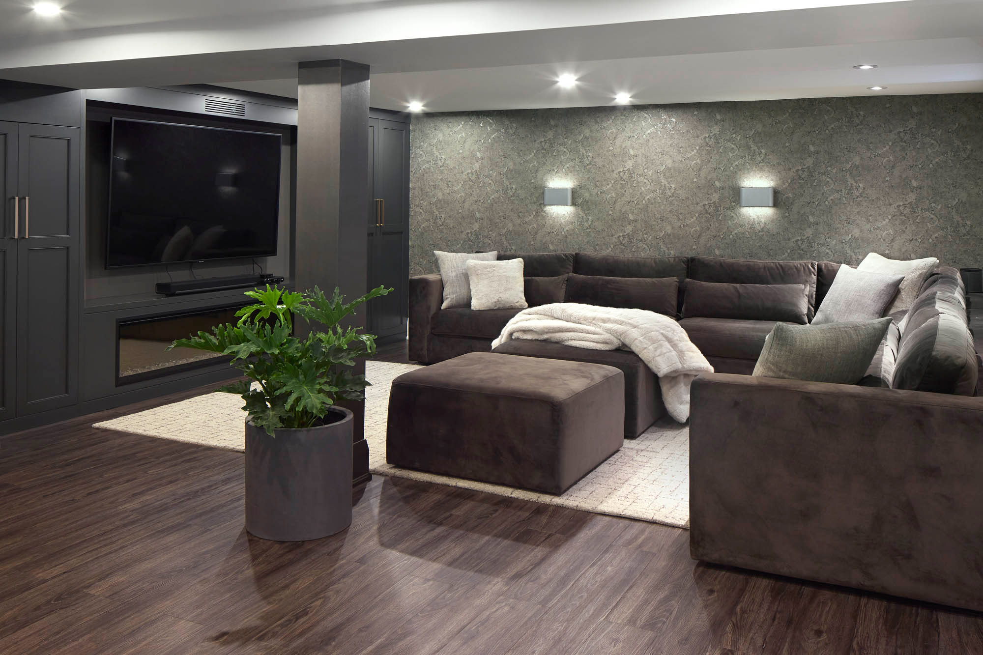 NUITH – FIFTY SHADES OF DIOR GRAY – BASEMENT MANCAVE THEATRE DESIGNED BY CELIA ALIDA RUTTE – PHOTOS BY DONNA GRIFFITH A large chocolate brown sectional with off white pillows, an off white area rug, and dark brown floors, in a theatre room with dark grey cabinets and a large TV.