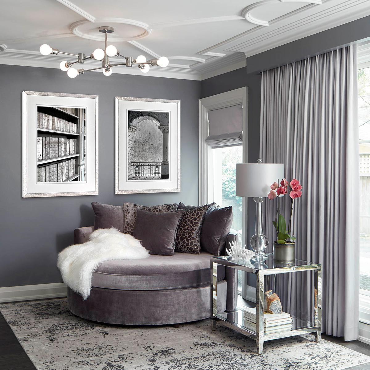 A reading nook designed in shades of purple grey with artwork on the walls, a large round reading chair, a side table, floor to ceiling drapery and an area rug.