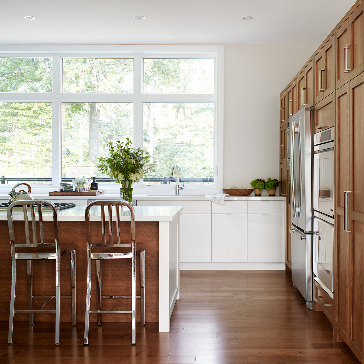 Two images of a modern kitchen designed with warm midtone woods, white countertops, high gloss white cabinets and a large picture window.