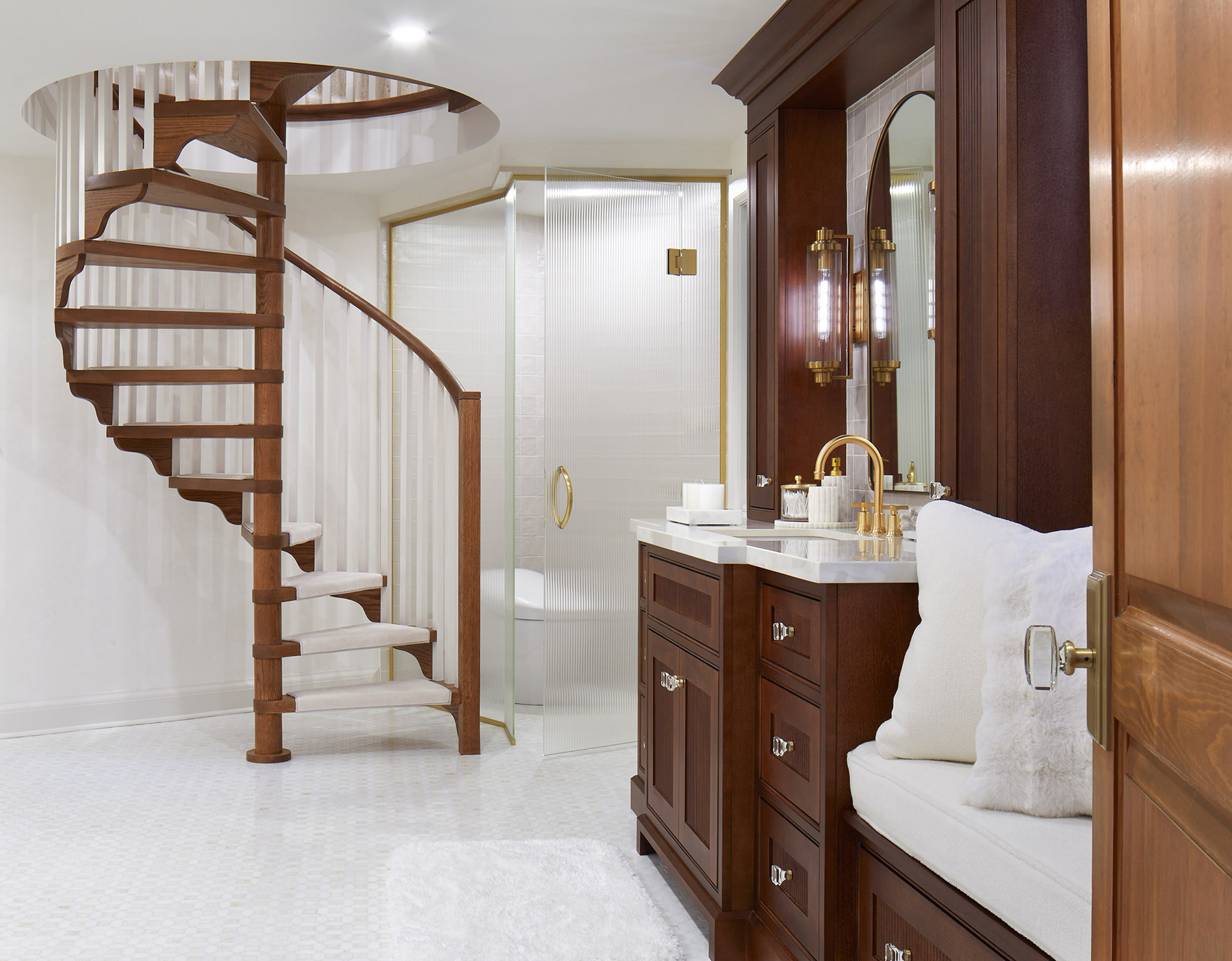 A photo of a bathroom showing a spiral staircase and the side view of a vanity in dark wood tones with light marble mosaic flooring and brass fixtures.