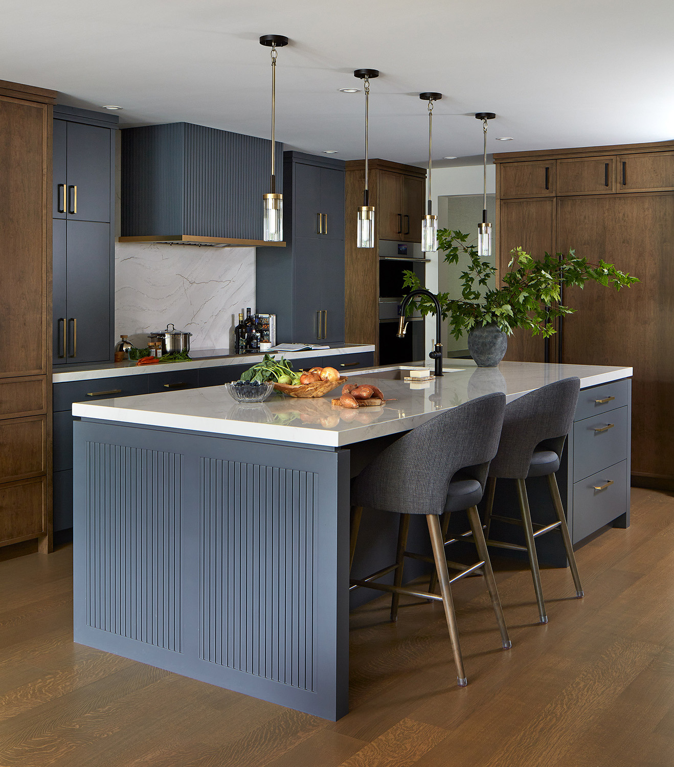 NUITH – STYLE AT HOME KITCHEN DESIGN – KITCHEN 01 DESIGNED BY CELIA ALIDA RUTTE – PHOTO BY DONNA GRIFFITH A kitchen photographed across a large island showcasing cabinetry in a combination of dark blue and warm caramel wood tones, brass and black accents and pendant lights.
