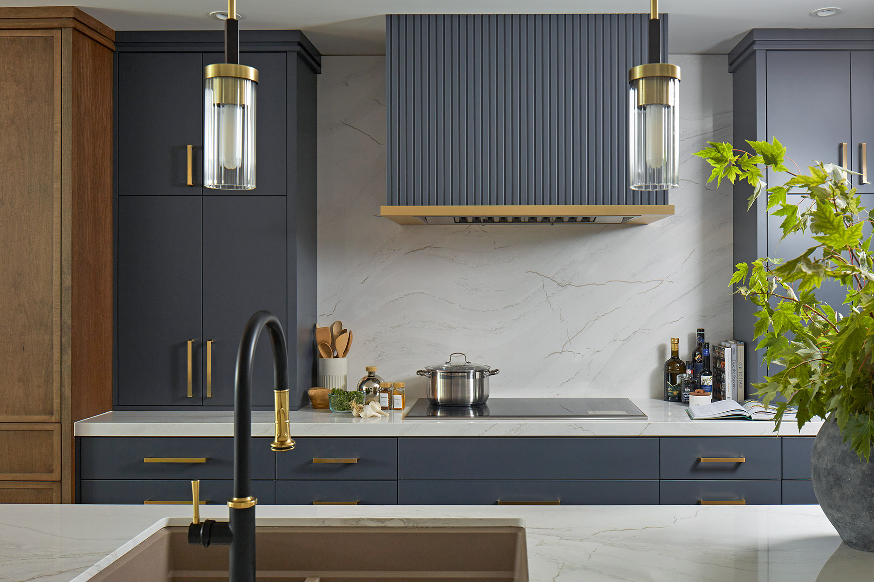 NUITH – STYLE AT HOME KITCHEN DESIGN – KITCHEN 02 DESIGNED BY CELIA ALIDA RUTTE – PHOTO BY DONNA GRIFFITH A kitchen photographed across a large island sink with black and brass faucet and pendant lights, and a mix of warm caramel wood tones with dark blue cabinetry.