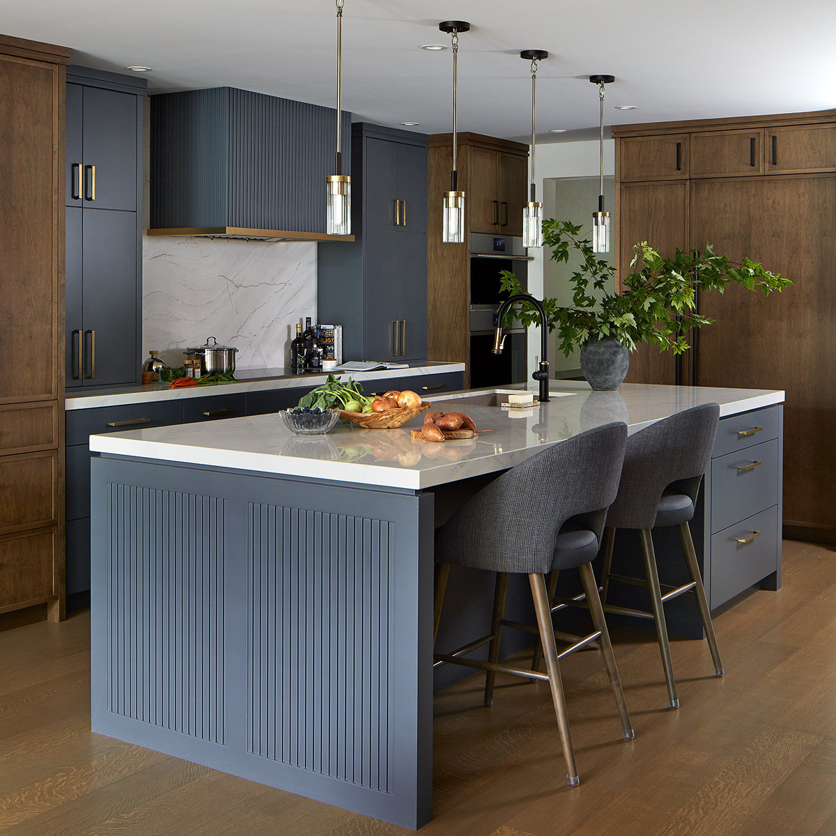 A kitchen photographed across a large island showcasing cabinetry in a combination of dark blue and warm caramel wood tones, brass and black accents and pendant lights.