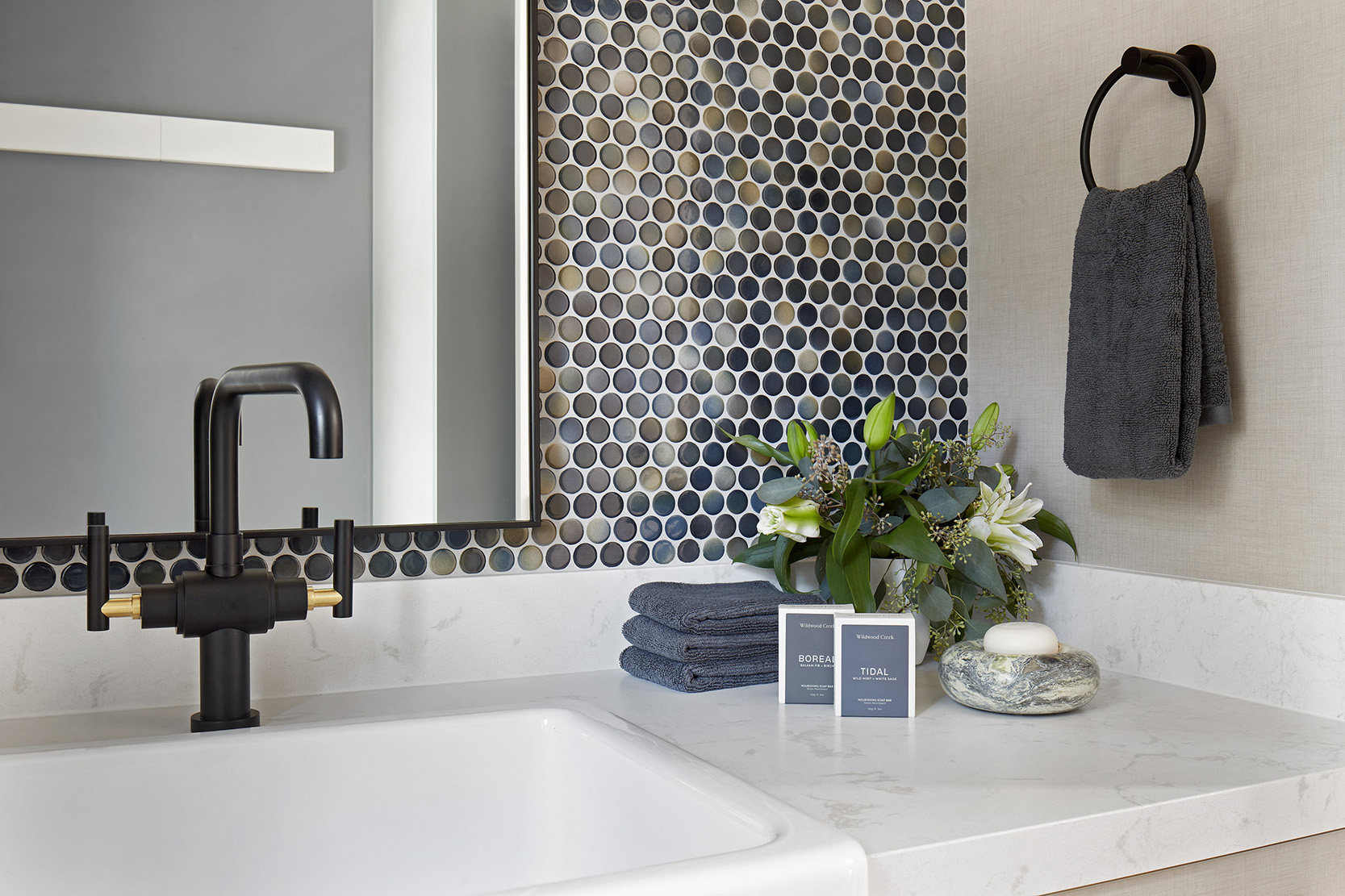 NUITH – ENLIGHTENED BY A TEMPEST – LAUNDRY POWDER ROOM DESIGNED BY CELIA ALIDA RUTTE – PHOTO BY DONNA GRIFFITH A close up image of the second floor laundry powder room showing the beautiful penny tile backsplash, matt black and brass faucet, and countertop accessories.