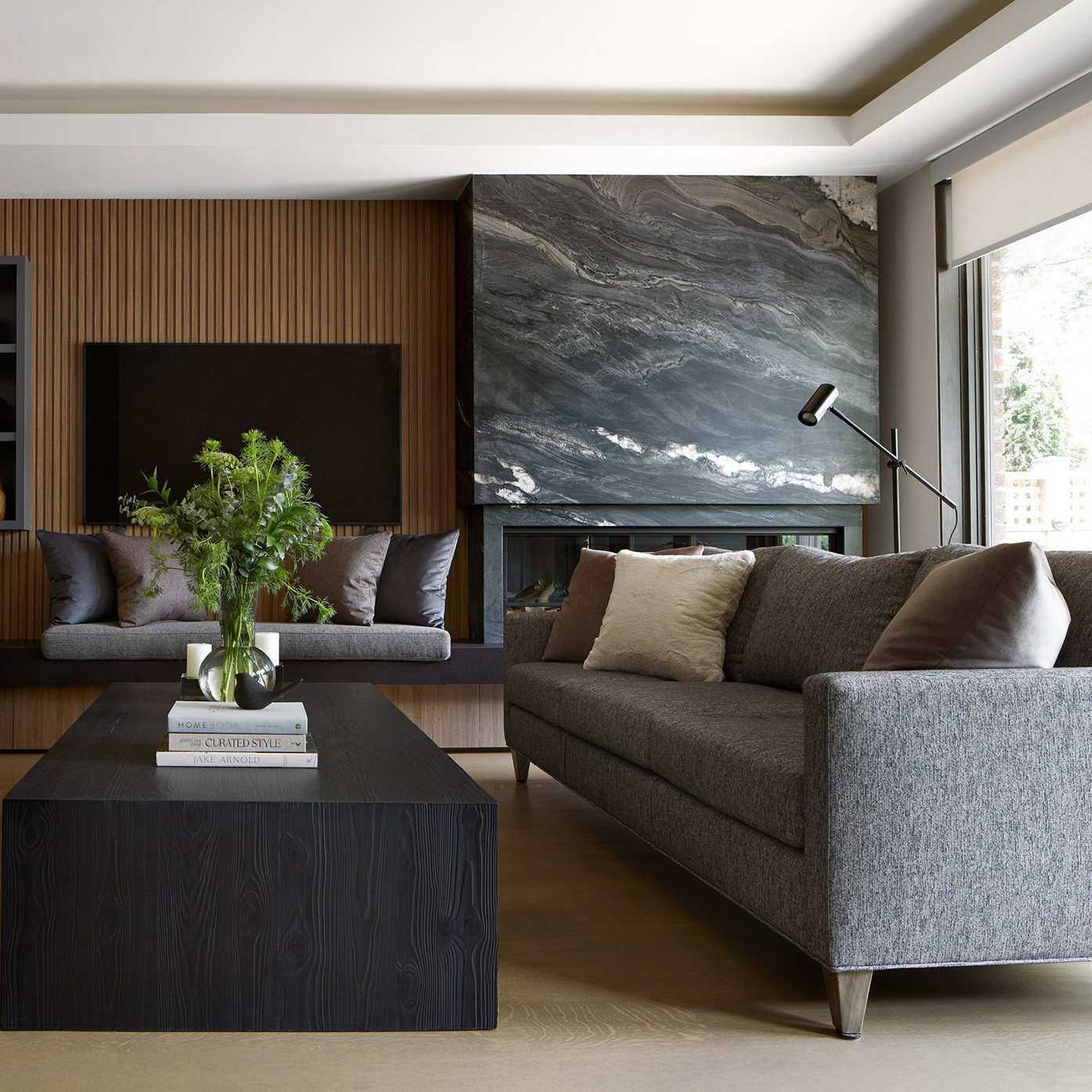 A living room designed in warm wood tones and soft natural gray blue accents with a stunning black tempest granite quartzite framing the fireplace.